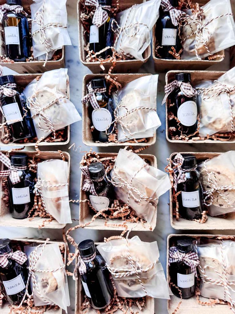 Nine gift boxes are shown from above, each containing a small bottle labeled "Vanilla," wrapped baked goods, and brown crinkle paper, tied with twine or gingham ribbon—thoughtful gifts perfect for pairing with simmer pot recipes.