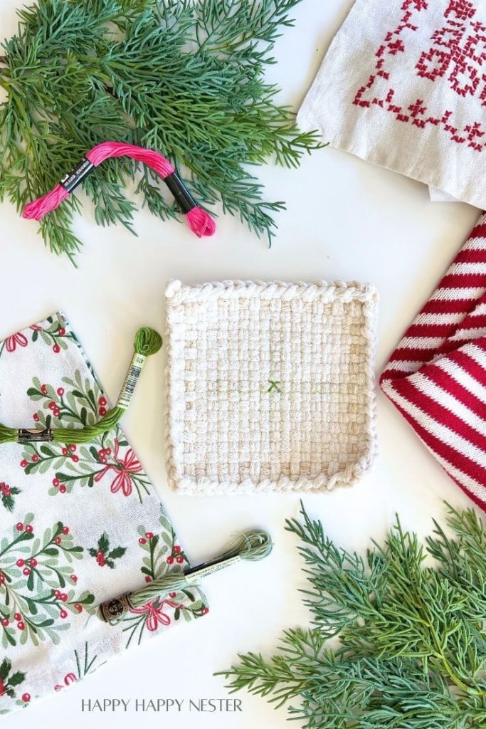 A crocheted square coaster is surrounded by green pine branches, embroidery floss in pink and green, and festive cloth napkins with holiday patterns, including red stripes and holly prints.