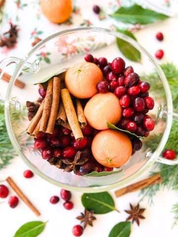 A clear glass bowl filled with fresh cranberries, whole clementines, cinnamon sticks, bay leaves, and star anise sits on a white surface—an inviting display perfect for simmer pot recipes for gifts. Scattered cranberries and spices complete the scene.