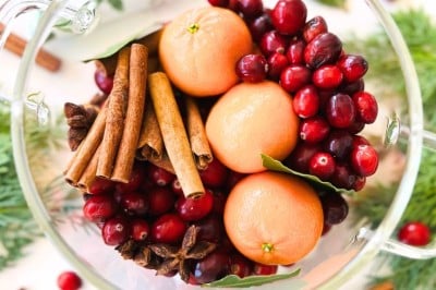 A clear glass bowl filled with fresh cranberries, whole clementines, cinnamon sticks, bay leaves, and star anise sits on a white surface—an inviting display perfect for simmer pot recipes for gifts. Scattered cranberries and spices complete the scene.
