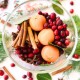 A clear glass bowl filled with fresh cranberries, whole clementines, cinnamon sticks, bay leaves, and star anise sits on a white surface—an inviting display perfect for simmer pot recipes for gifts. Scattered cranberries and spices complete the scene.