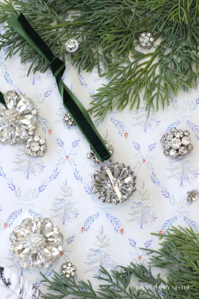 Vintage rhinestone brooches and a jeweled pendant with a green velvet ribbon are arranged on a wintry patterned background, surrounded by pine branches and evergreen sprigs.