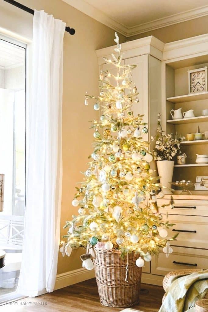 A slim Christmas tree in a woven basket, adorned with white and pastel ornaments, glowing lights, and charming Christmas crafts, stands beside a window with sheer curtains in a cozy, beige-toned living room.