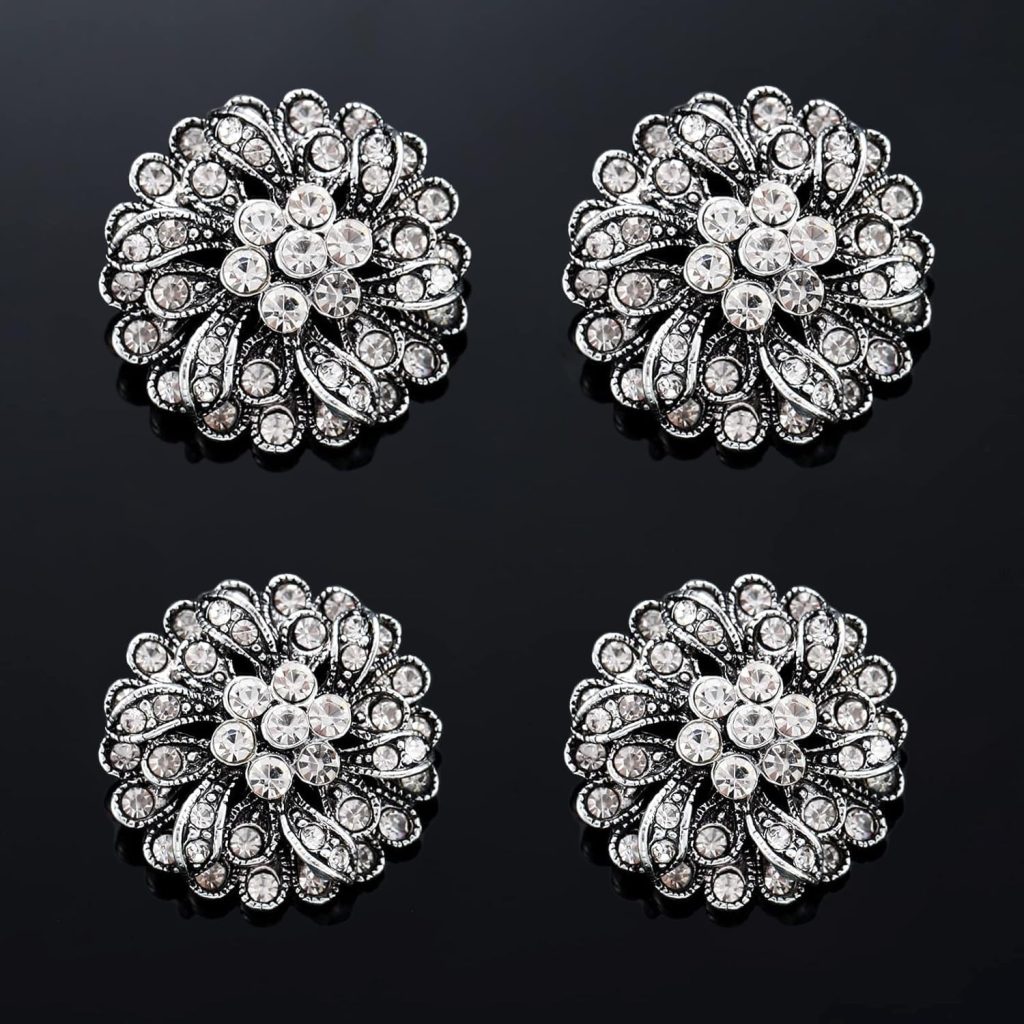 Four ornate, round, silver brooches with intricate floral designs and sparkling clear rhinestones evoke vintage style ornaments, beautifully arranged in a grid on a glossy black background.
