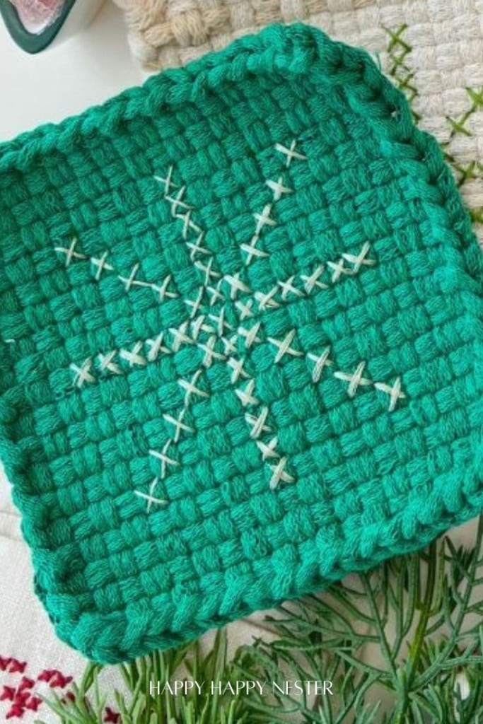 A green woven square potholder features a large white embroidered snowflake design in the center, displayed on a textured background with other fabric items partially visible.