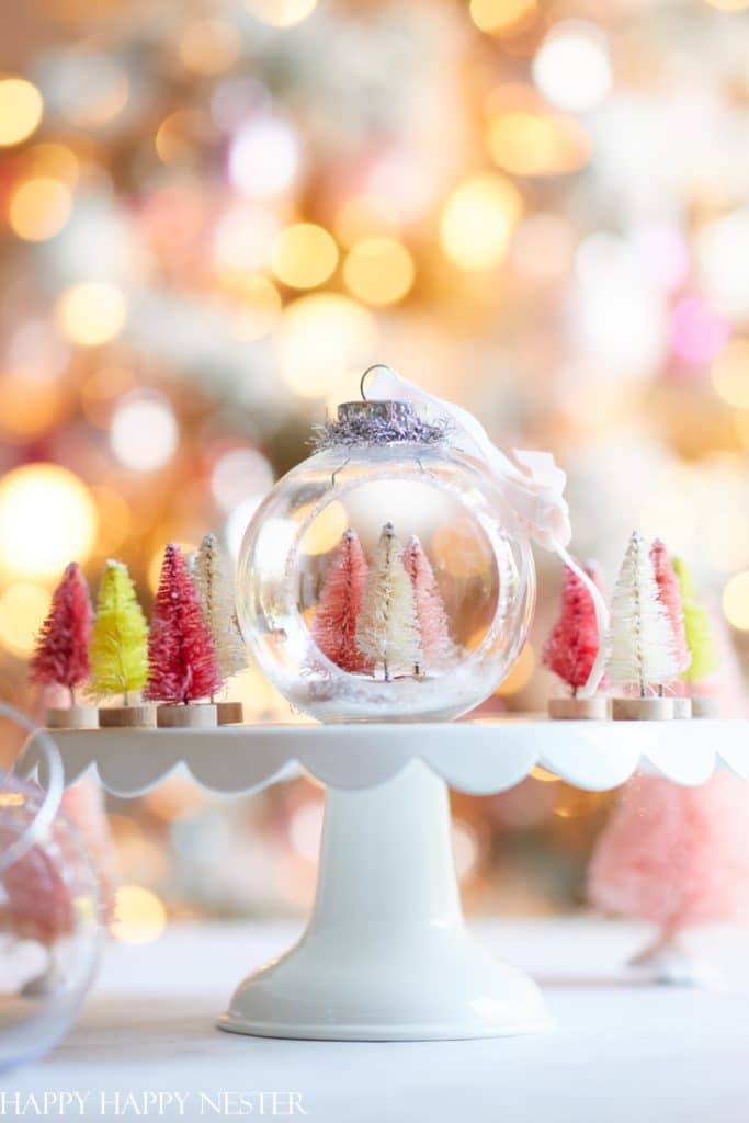 A clear glass ornament filled with tiny pink and white bottle brush trees sits on a white cake stand, surrounded by more miniature trees—an enchanting display for Christmas crafts. The background features warm, out-of-focus Christmas lights.