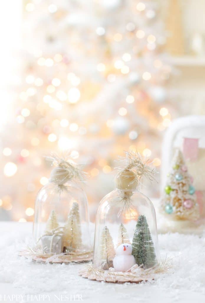 Two glass domes with miniature Christmas trees and a snowman inside sit on a snowy surface, showcasing charming Christmas tree crafts against a blurred, light-filled holiday backdrop for a festive and cozy scene.