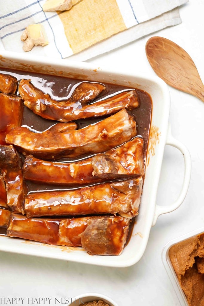 Raw pork ribs coated in a dark brown marinade sit arranged in a white rectangular baking dish, ready to become a savory Thanksgiving food. A wooden spoon, yellow and white towel, and bowls of brown sugar and ginger are nearby.