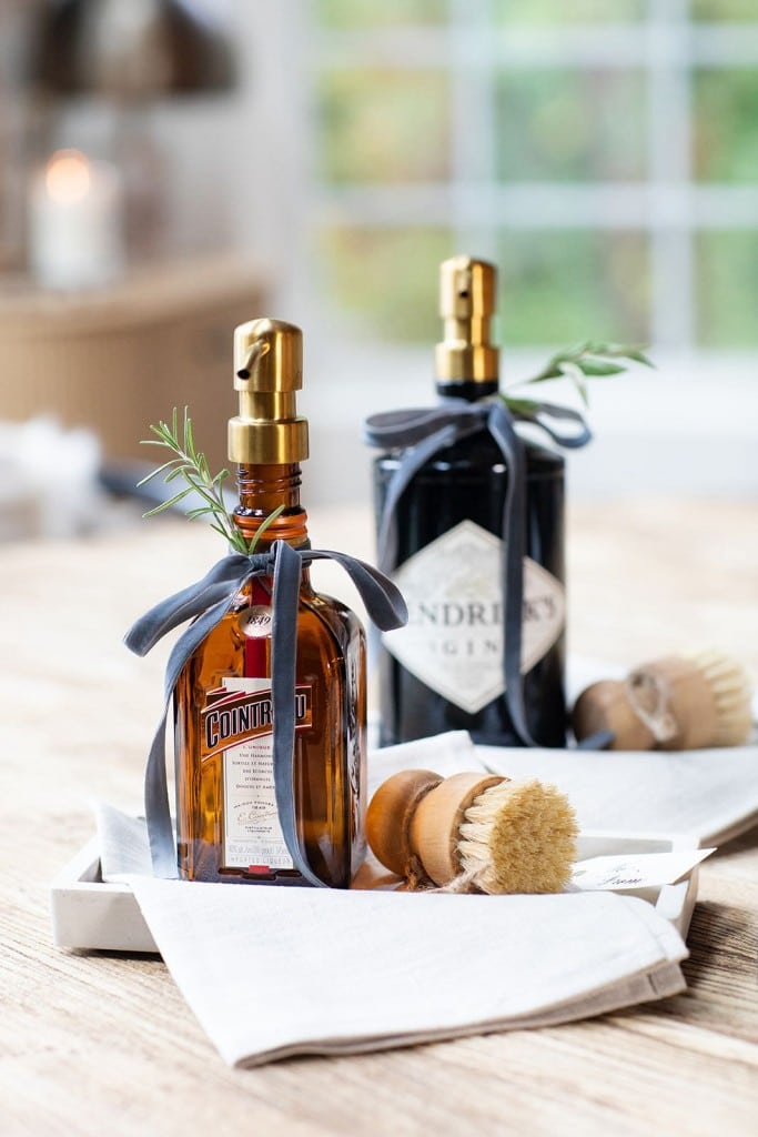 Two upcycled liquor bottles with soap dispensers, perfect as unique gifts, one brown and one black, are decorated with bows and rosemary sprigs. They sit on a tray with two scrub brushes and a folded cloth on a wooden table.