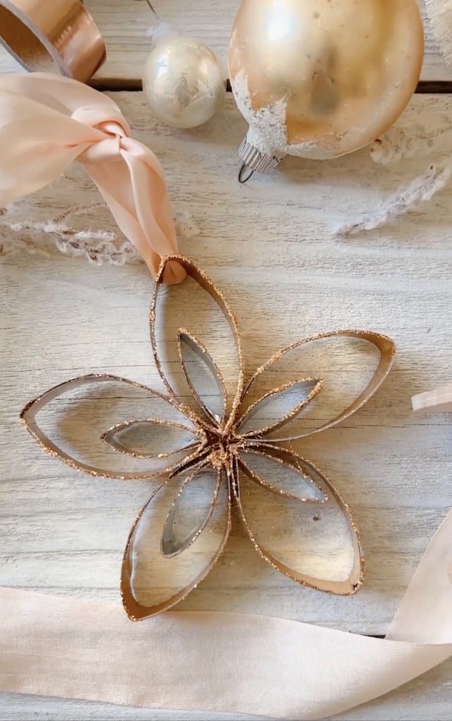 A handmade ornament shaped like a flower, crafted from cut toilet paper rolls and dusted with gold glitter, lies on a white wooden surface—perfect for Christmas tree crafts—next to pink ribbon and gold Christmas baubles.