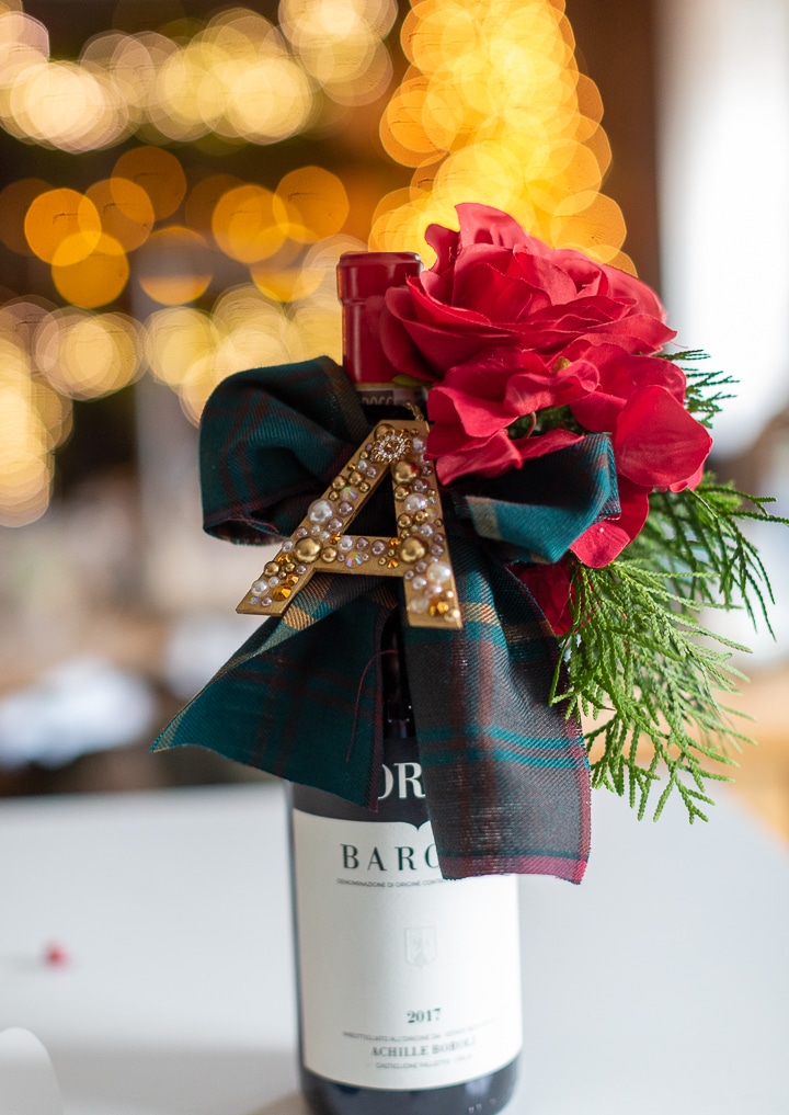 A wine bottle decorated with a large red flower, green foliage, a green plaid ribbon, and a gold letter "A" adorned with pearls—perfect for Simmer Pot Gifts—set against a background of warm, blurry string lights.