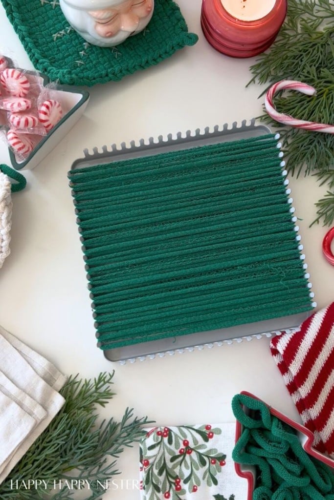 A green potholder is being woven on a square loom, surrounded by Christmas decorations, peppermint candies, candy canes, candles, greenery, and festive linens—perfect for DIY Christmas gifts on a white surface.