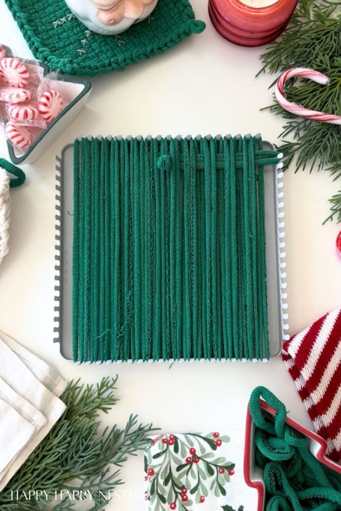 A green woven potholder in progress sits in a metal loom, surrounded by festive holiday decor, greenery, candy, and red-and-white textiles on a white surface.