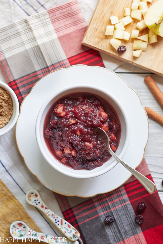 A bowl of chunky cranberry sauce with apple pieces and a spoon, placed on a white plate. Nearby are chopped apples, brown sugar, cinnamon sticks, and measuring spoons on a plaid tablecloth—perfect thanksgiving food.