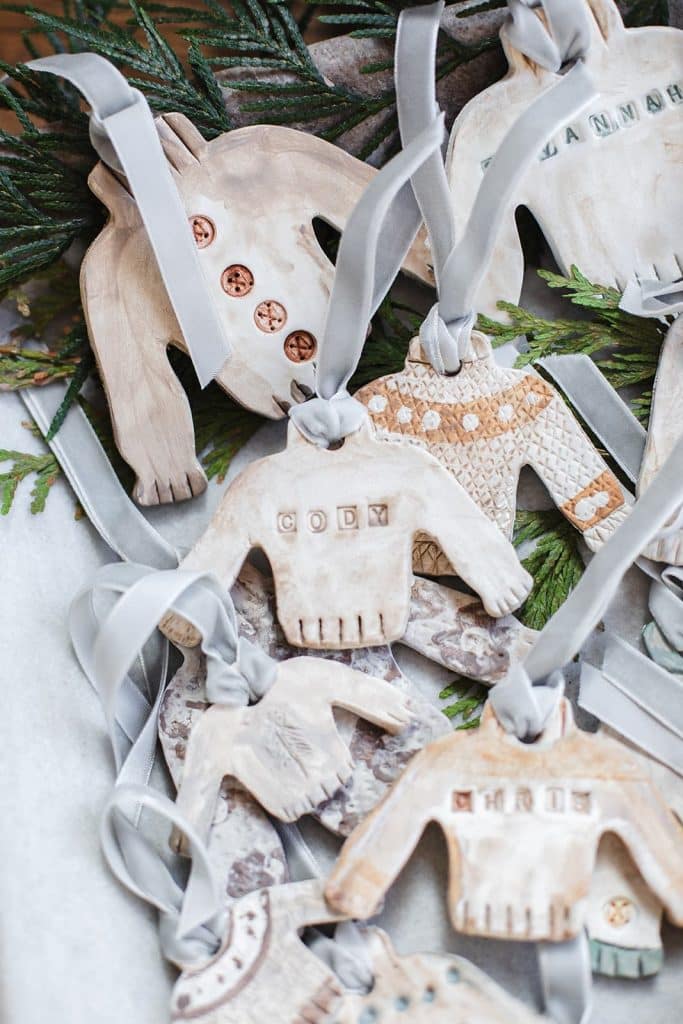 A collection of handmade clay DIY Christmas ornaments shaped like sweaters, decorated with different patterns and names, tied with gray ribbons, displayed on green pine branches.