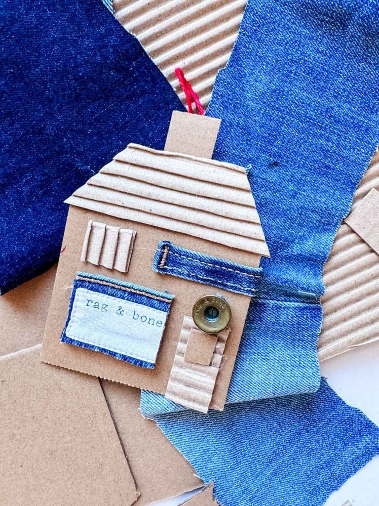A small house craft made from cardboard and denim fabric scraps, with a button for a doorknob, labeled "rag & bone." Surrounded by more fabric and cardboard pieces, it evokes the charm of vintage style ornaments perfect for DIY Christmas ornaments.
