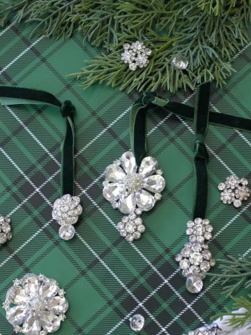 Three sparkling, vintage style Christmas ornaments with crystal designs hang from green velvet ribbons on plaid wrapping paper, surrounded by pine branches and additional brooches—perfect inspiration for a festive DIY display.