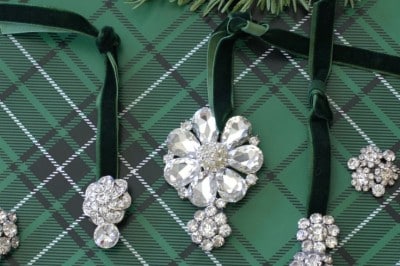 Three sparkling, vintage style Christmas ornaments with crystal designs hang from green velvet ribbons on plaid wrapping paper, surrounded by pine branches and additional brooches—perfect inspiration for a festive DIY display.