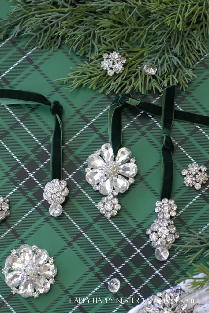 Three sparkly vintage brooches hang from green velvet ribbons atop green plaid fabric, surrounded by evergreen branches and more crystal brooches—perfect inspiration for festive Christmas crafts.
