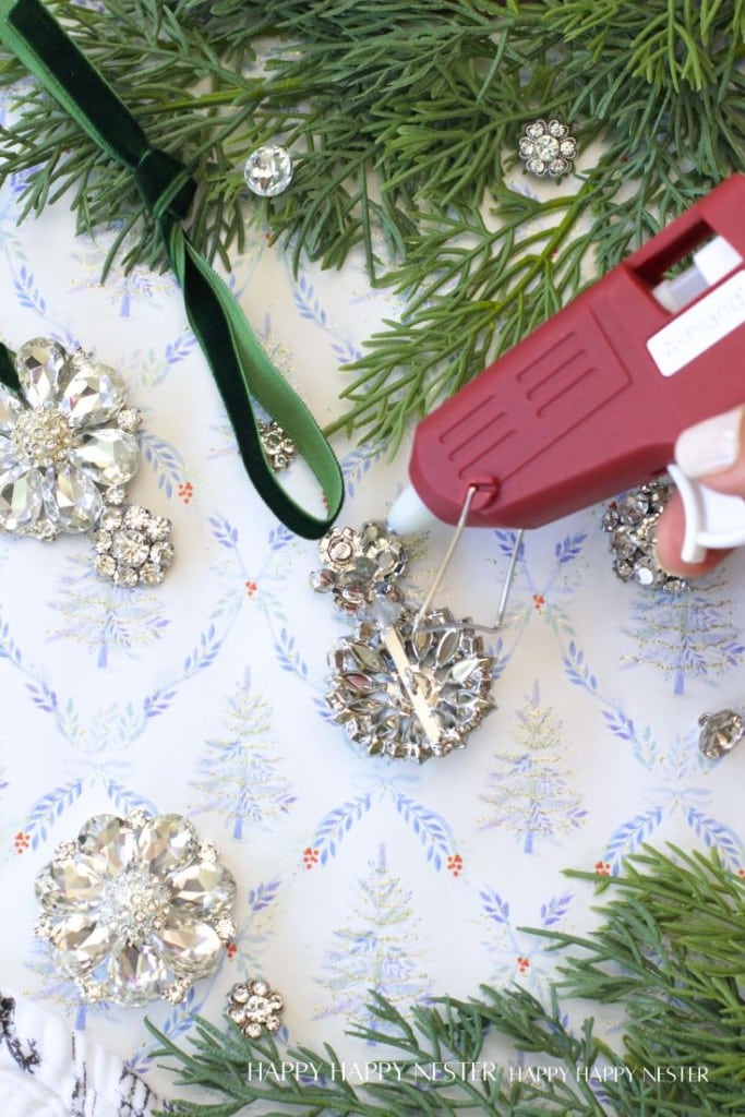 A hand uses a hot glue gun to attach a green velvet ribbon to a sparkly jeweled ornament, surrounded by greenery and other jeweled decorations on a festive background with pine tree illustrations.