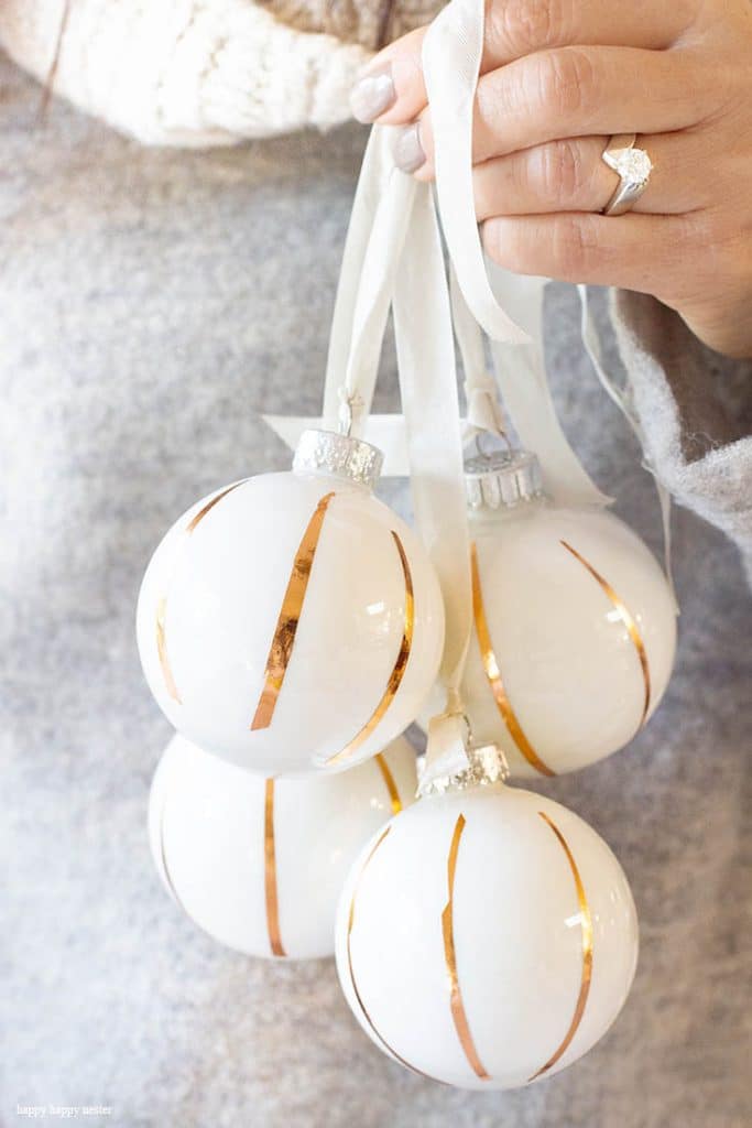 A hand with a diamond ring holds four white Christmas ornaments decorated with thin gold diagonal stripes and white ribbons—perfect for Christmas tree crafts. The person is wearing a cozy, light gray sweater.