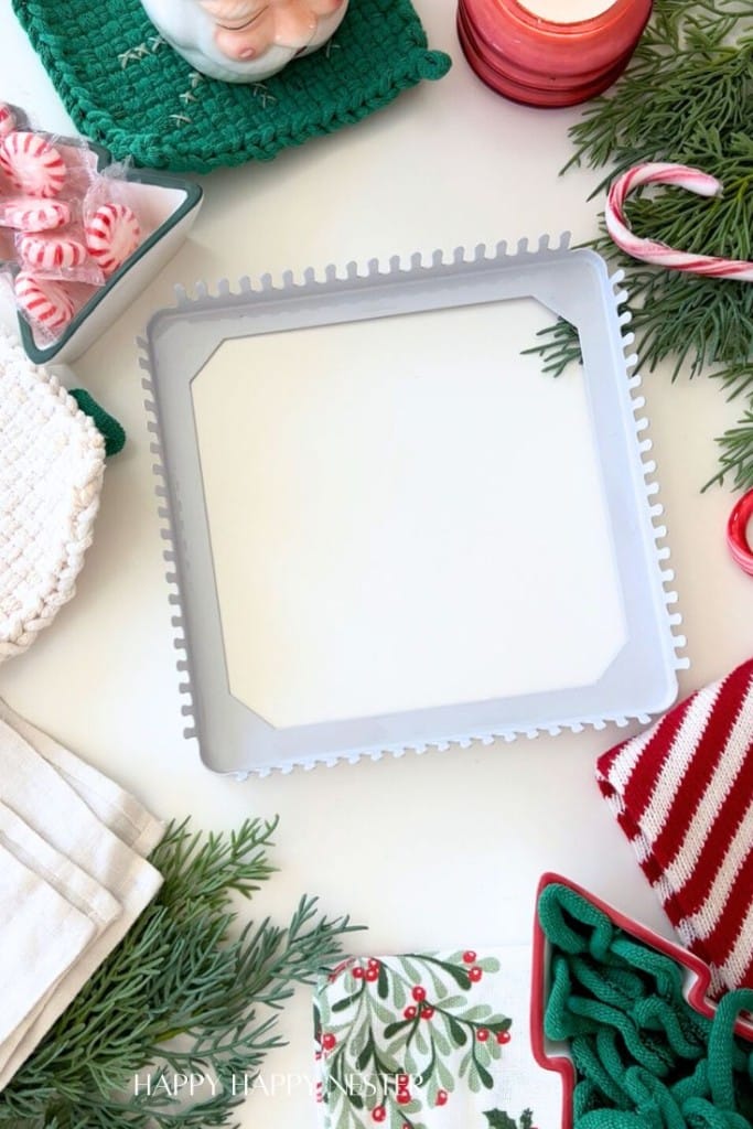 A square white loom sits on a festive table surrounded by holiday decor, candy canes, greenery, a candle, striped and green potholders, and napkins, creating a cheerful Christmas crafting scene.