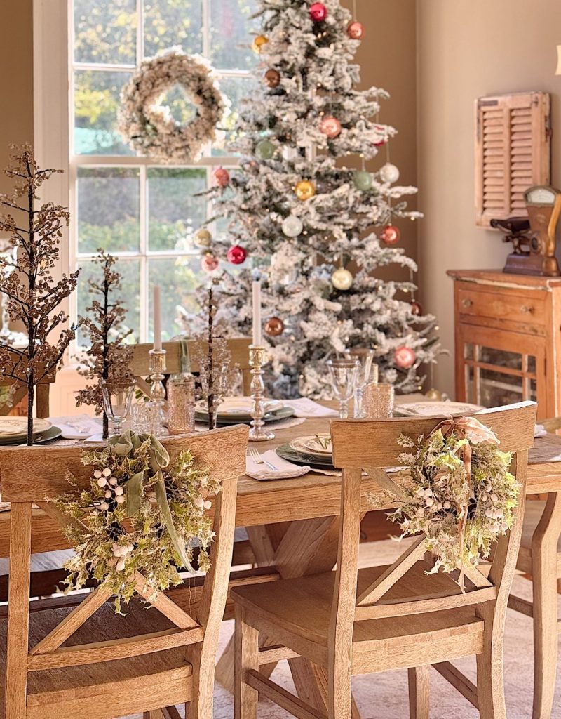 A festive dining room with a wooden table set for a meal, decorated with greenery and glassware. My favorite Christmas decor—a flocked tree adorned with ornaments—stands behind the table, while sunlight streams through a window crowned by a wreath.