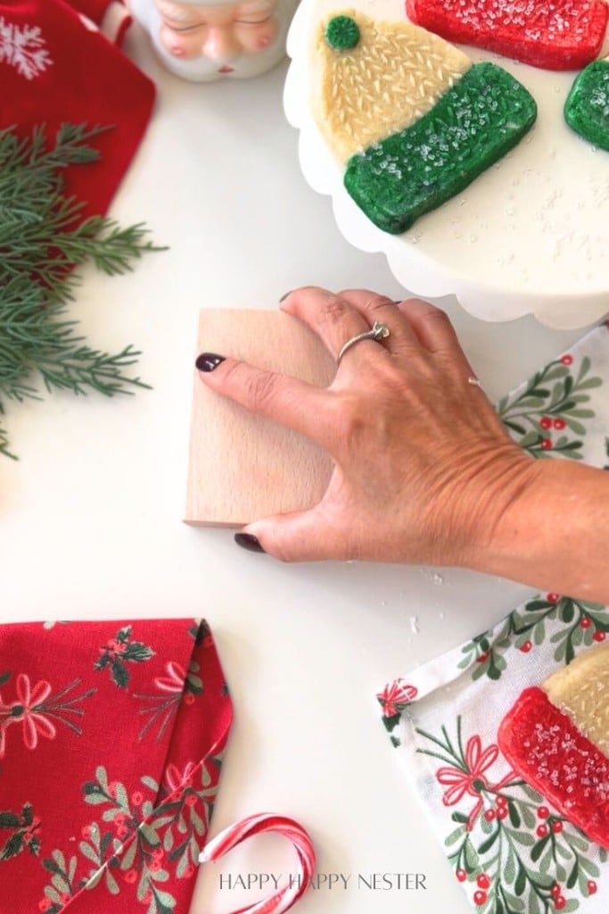 A hand with dark nail polish holds a small wooden block on a white table decorated with holiday items, including festive cookies, greenery, a Santa mug, red napkins, and a candy cane.