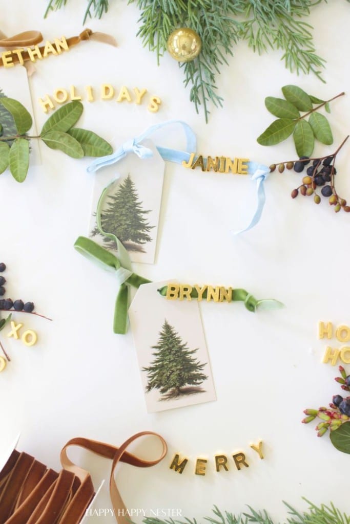 DIY Christmas gift tags featuring evergreen tree illustrations are tied with ribbon and adorned with gold alphabet letters spelling names like "JANINE" and "BRYNN." Sprigs of greenery and gold letters create festive words like "HOLIDAYS" on a white surface.