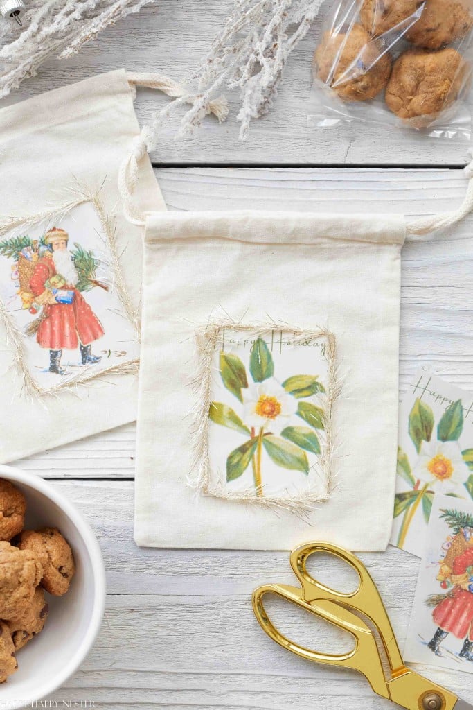 A drawstring gift bag with a painted flower tag sits on a white wooden surface, surrounded by cookies, a bowl, gold scissors, another decorated bag, tags, and frosted branches—my favorite gift wrap inspiration for the season.