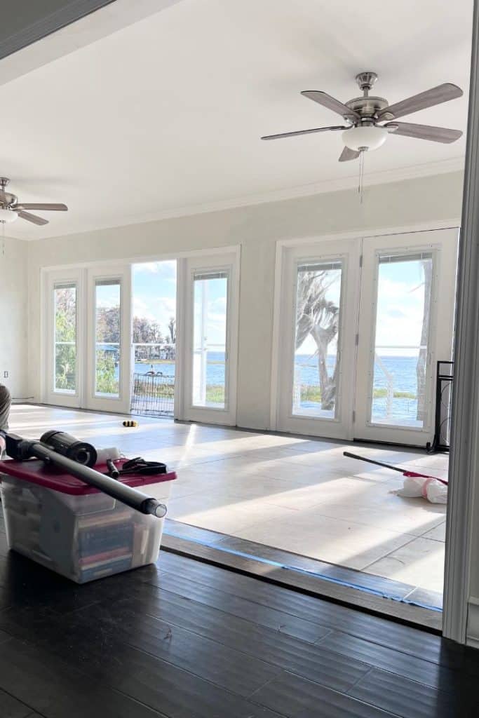 Sunlit room with large windows and glass doors overlooking a waterfront, ceiling fans above, and cleaning supplies in a storage bin on a dark wooden floor—perfect for finding my favorite gift wrap inspiration.