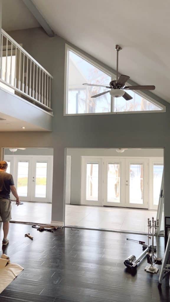 A person stands in a spacious, modern living room with high ceilings, large windows, and double doors letting in natural light. Nearby, some renovation tools hint at a DIY project—perhaps inspired by my favorite gift wrap inspiration.