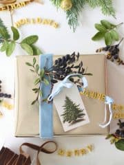 DIY Christmas Gift Tags A gold-wrapped gift with a blue ribbon, botanical sprig, and DIY Christmas Gift Tag sits on a white surface. Surrounding it are greenery, berries, and yellow letter cutouts spelling festive words like "HOLIDAYS," "MERRY," and "HO HO HO.