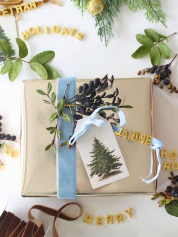 A gold-wrapped gift with a blue ribbon, botanical sprig, and DIY Christmas Gift Tag sits on a white surface. Surrounding it are greenery, berries, and yellow letter cutouts spelling festive words like "HOLIDAYS," "MERRY," and "HO HO HO.