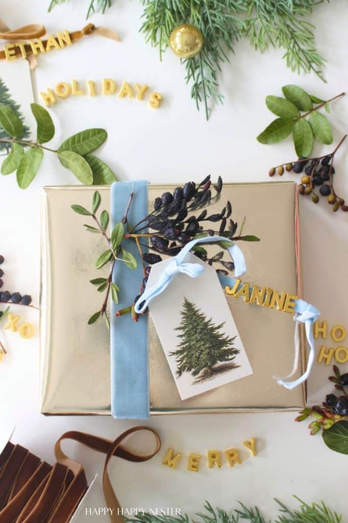 A gold-wrapped gift with a blue ribbon, botanical sprig, and DIY Christmas Gift Tag sits on a white surface. Surrounding it are greenery, berries, and yellow letter cutouts spelling festive words like "HOLIDAYS," "MERRY," and "HO HO HO.