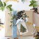 A gold-wrapped gift with a blue ribbon, botanical sprig, and DIY Christmas Gift Tag sits on a white surface. Surrounding it are greenery, berries, and yellow letter cutouts spelling festive words like "HOLIDAYS," "MERRY," and "HO HO HO.