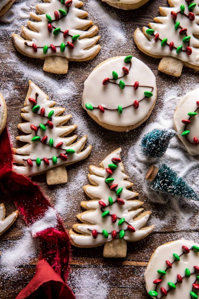 Iced Christmas cookies—my favorite Christmas cookies—are shaped like trees and circles, decorated with white icing and red and green sprinkles on a wooden surface dusted with powdered sugar, surrounded by mini snow-covered trees and a red ribbon.