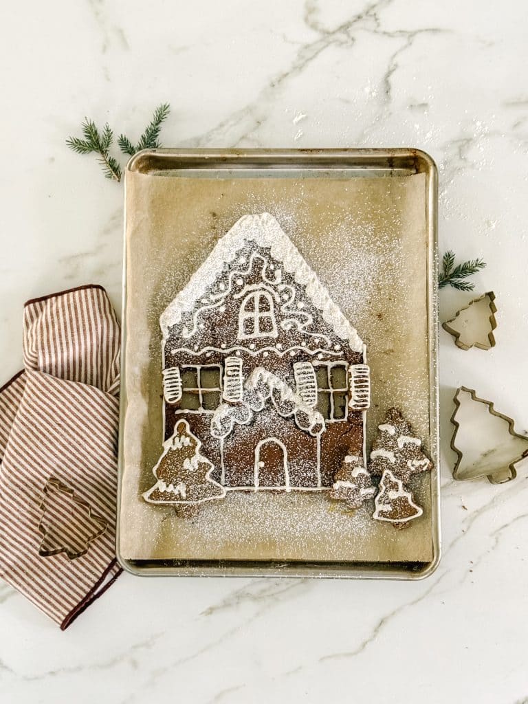 A decorated gingerbread house cookie dusted with powdered sugar sits on a baking sheet beside tree-shaped cookies, striped oven mitts, and tree-shaped cookie cutters—perfect inspiration for any Molded Cookies Recipe. Pine sprigs accent the scene.