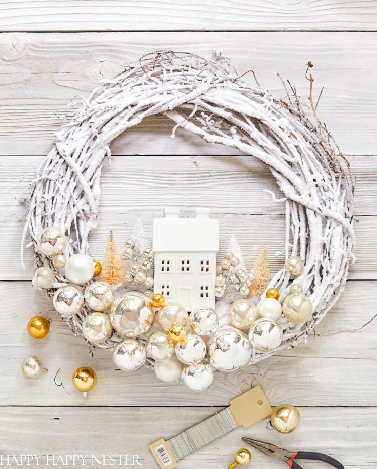 A white twig wreath with gold and silver ornaments, golden trees, and a small white house sits on a white wooden surface—my favorite Christmas decor. Crafting supplies lie nearby at the bottom right.