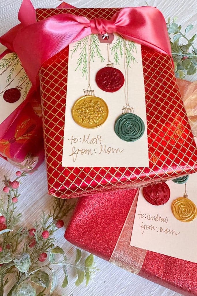 Two Christmas gifts wrapped in red paper with pink ribbons and DIY gift tags decorated with wax seals and festive designs, addressed from "mom" to "Matt" and "Andrew." Lush greenery adds a holiday touch in the background.