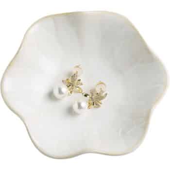 A pair of gold earrings with leaf designs and white pearl drops rest in a scalloped, white ceramic dish.