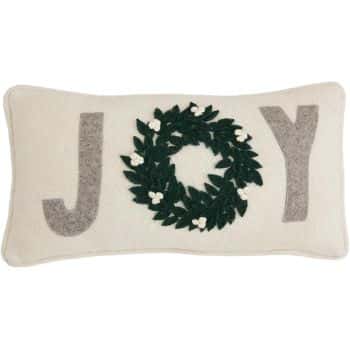 A rectangular cream-colored pillow with the word "JOY" on it. The "O" is replaced by a green wreath with small white flowers, while the "J" and "Y" are made of gray fabric.