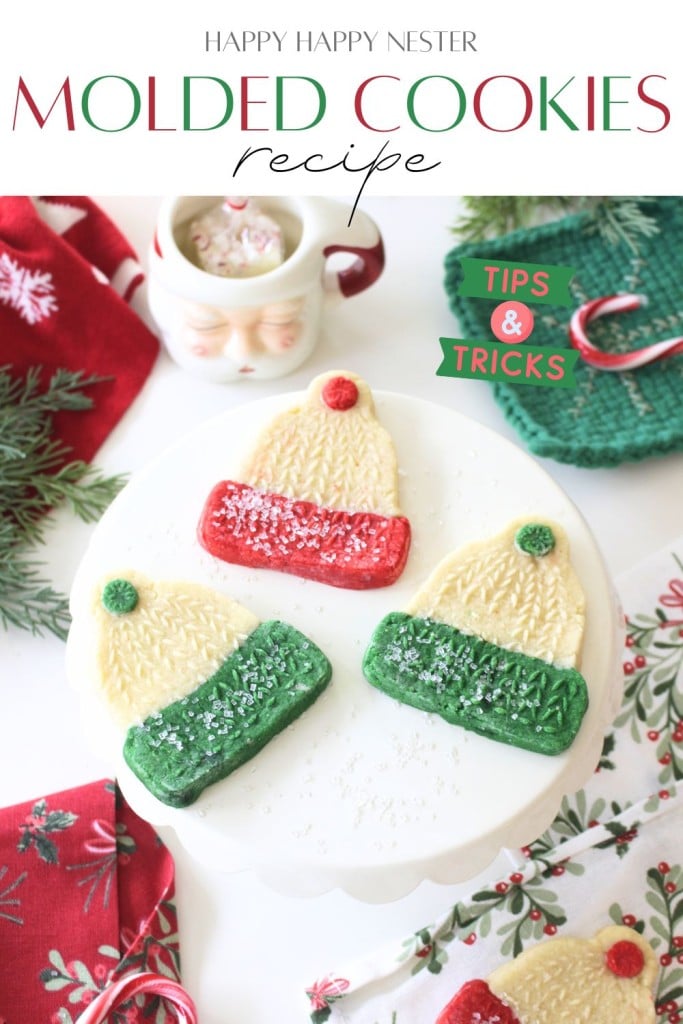 Festive molded cookies shaped like winter hats, decorated in red, green, and white, are displayed on a white cake stand with holiday decor and a Santa mug in the background. Text overlay reads “Molded Cookies Recipe.”.