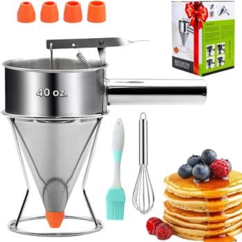 A stainless steel 40 oz. batter dispenser with a handle and orange nozzle, four extra nozzles, a small whisk, a silicone basting brush, a box, and a stack of pancakes topped with berries.
