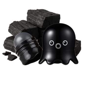 A cute, black, octopus-shaped container with a charcoal facial balm sits open next to black charcoal pieces. The container has big, round eyes and the balm is partially exposed.