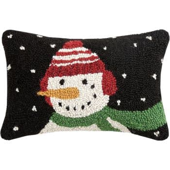 Rectangular black pillow featuring a hooked design of a smiling snowman with a red hat, red earmuffs, green scarf, orange carrot nose, and white snowflakes in the background.