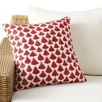 A white couch with a wicker armrest features a square pillow decorated with a pattern of red Christmas trees on a white background.
