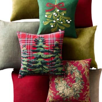 A group of decorative pillows in festive colors and patterns, including plaid with pine trees, embroidered mistletoe, olive green, solid red, beige, and a pillow with a floral wreath design.