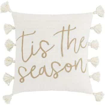 A white decorative pillow with beige tassels on the edges and the phrase "'tis the season" written in large, cursive gold lettering across the front.