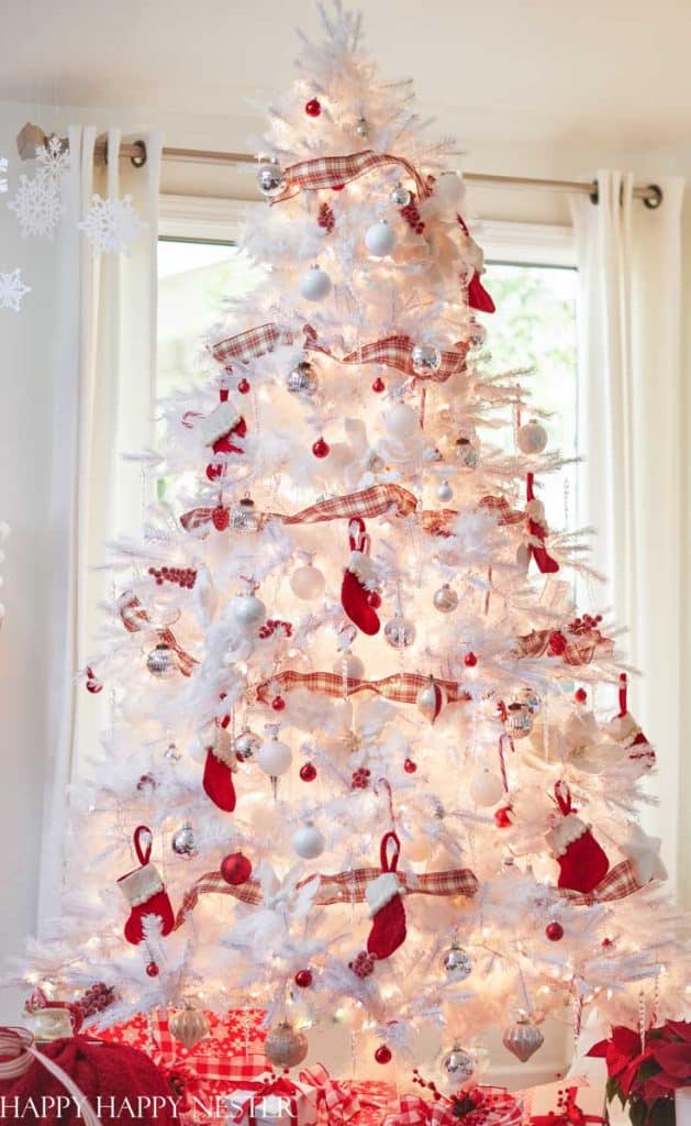 A white Christmas tree decorated with red and white ornaments, plaid ribbon, red mittens, and white lights—my favorite Christmas decor—stands in front of bright windows with white curtains and wrapped presents underneath.